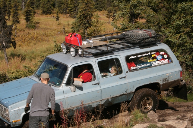 Light blue SUV with roof rack, spare tire, and gas cans on rough terrain