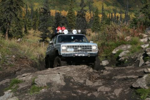 1990 Chevrolet Suburban - A silver SUV navigates a challenging off-road trail with a rocky and muddy terrain.