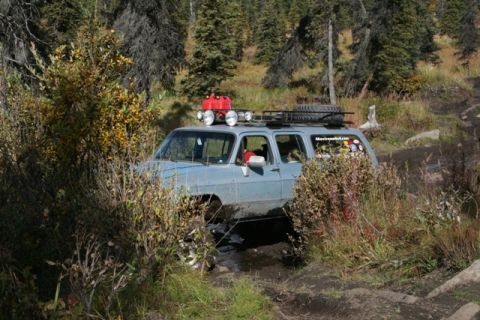 1990 Chevrolet Suburban - This modified SUV is tackling a challenging off-road trail surrounded by trees and foliage.