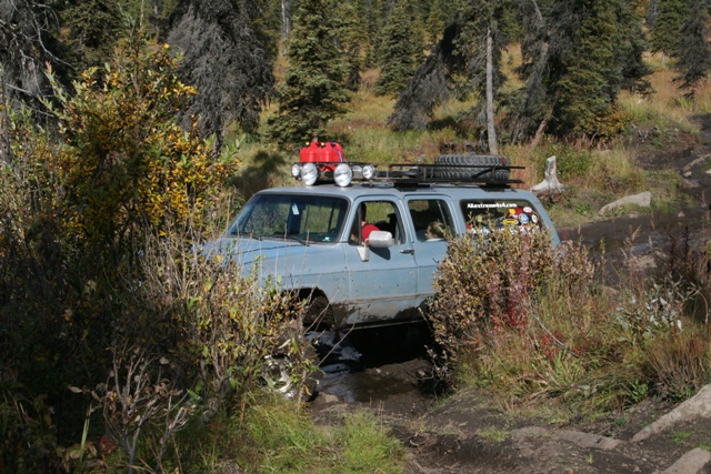 Blue SUV with roof rack, spare tire, and gas cans navigating a muddy trail