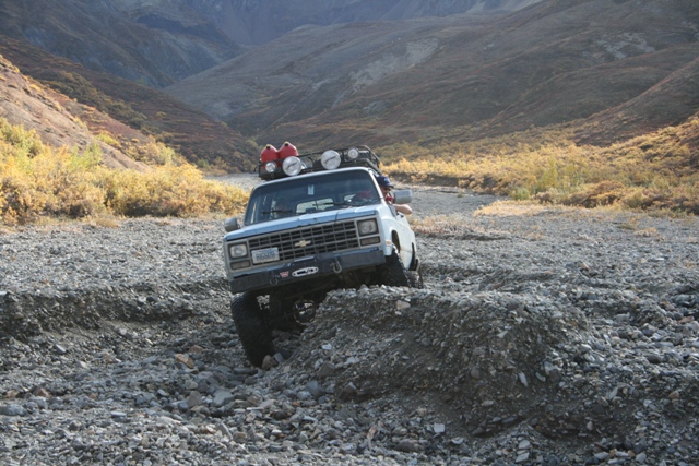 Pickup truck driving over rocks in a dry creek bed with mountains in background. Has extra lights and gas cans on roof
