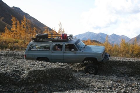 1990 Chevrolet Suburban - An older model SUV navigates a rocky, off-road path with fall foliage and mountains visible.