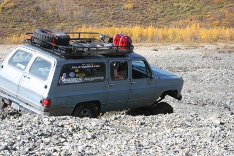 1990 Chevrolet Suburban - A blue 1990 Chevrolet Suburban navigates challenging rocky terrain, equipped with a roof rack carrying a spare tire and fuel cans.