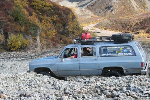 1990 Chevrolet Suburban - A light blue Chevrolet Suburban navigates a rocky creek bed with autumn foliage in the background.