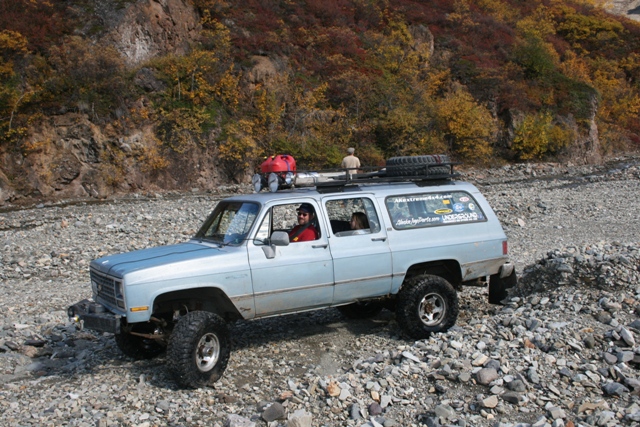 1990 Chevrolet Suburban with roof rack and spare tire, driving on a rocky riverbed