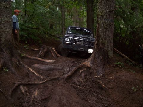 1985 Toyota Pickup/SR5 - A person watches an off-road vehicle crawl over roots and mud on a challenging forest trail.