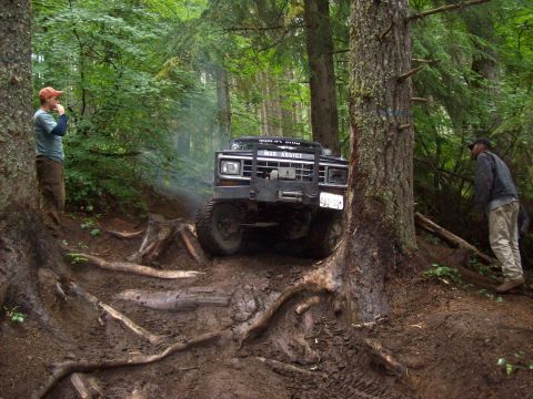 1985 Toyota Pickup/SR5 - A black off-road truck maneuvers through a challenging, muddy forest trail with exposed roots.