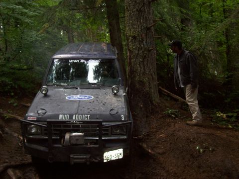 1985 Toyota Pickup/SR5 - A Ford pickup truck, modified for off-road use with "MUD ADDICT" on its hood, is seen parked on a muddy trail surrounded by trees.