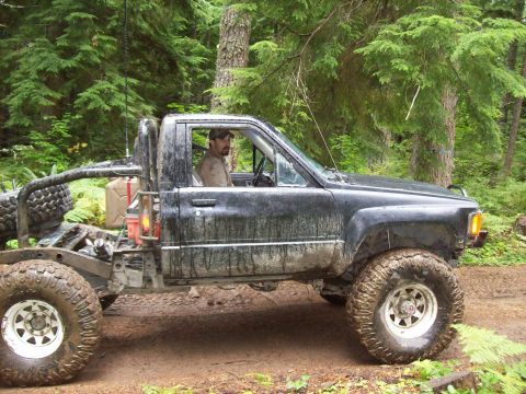 1985 Toyota Pickup/SR5 - A man drives a heavily modified Toyota Pickup through a wooded area, equipped with large tires and extra fuel cans.