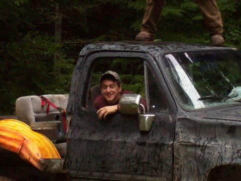 1985 Toyota Pickup/SR5 - A man smiles from the driver's seat of a mud-covered truck, while another person stands on the roof.