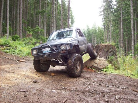 1985 Toyota Pickup/SR5 - A modified black Toyota pickup truck navigates a rocky trail in a forest setting.