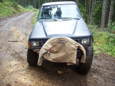 1985 Toyota Pickup/SR5 - A black Toyota Pickup truck is parked on a dirt road in a forest, with a jacket draped over its front.
