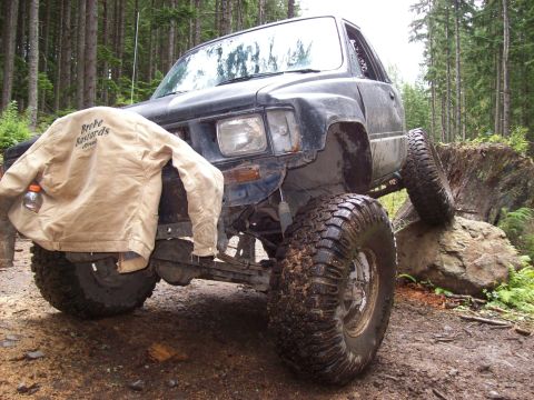 1985 Toyota Pickup/SR5 - A black 1985 Toyota Pickup SR5 is shown with large off-road tires, navigating rocky terrain in a forest.