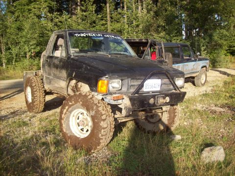 1985 Toyota Pickup/SR5 - Two off-road vehicles are parked on a dirt path surrounded by trees.