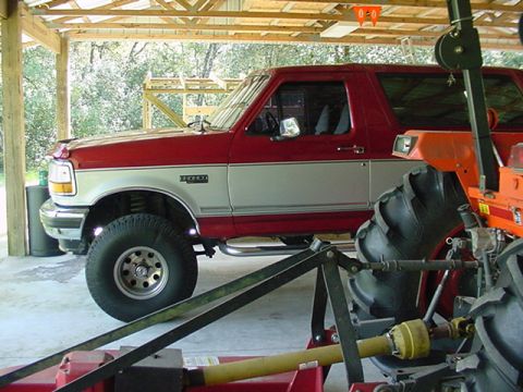 1996 Ford Bronco - A red and silver Ford Bronco is parked under a covered area next to farm equipment.
