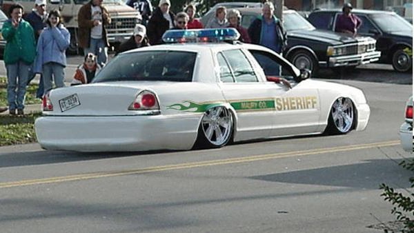 Lowered white Sheriff sedan with chrome wheels and light bar, viewed from rear side on a street
