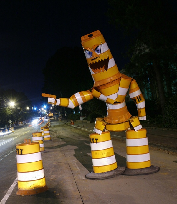 Angry monster figure built from orange-and-white striped traffic barrels on a road at night