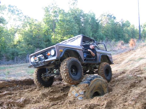 1967 Ford Mustang - A driver maneuvers a lifted Ford Bronco through challenging muddy terrain, with one wheel caught on an obstacle.