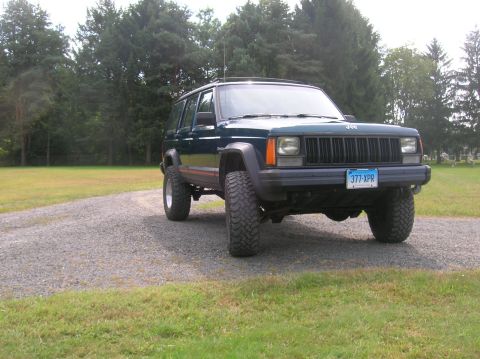 1995 Jeep Cherokee - A dark green Jeep Cherokee is shown from a 3/4 front angle on a gravel road with trees in the background.