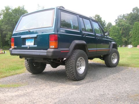 1995 Jeep Cherokee - This dark green Jeep Cherokee XJ is shown from the rear, parked on a gravel surface with a grassy area and trees in the background.