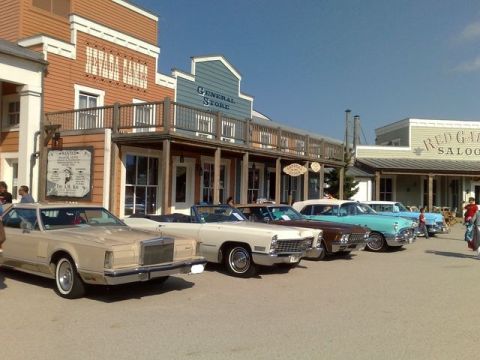 1991 Ford LTD Crown Victoria - A row of vintage American cars are displayed in front of a western-themed town facade.