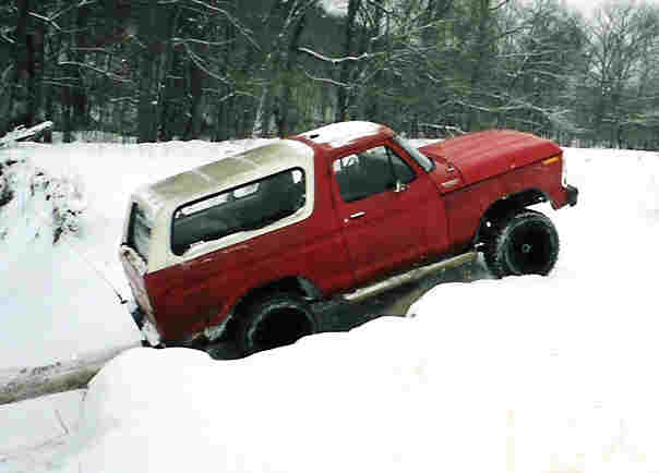 Red and white 1978 Ford Bronco partially submerged in a snow-filled ditch, with trees in the background