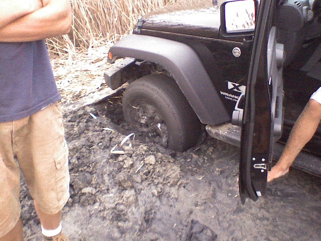 Black Jeep Wrangler stuck in mud, front wheel deep, door open, person standing nearby