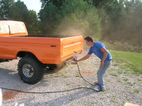 1975 Ford F-250 - A person spray paints the rear quarter panel of an orange 1975 Ford F-250 pickup truck.