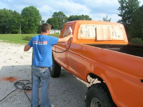 1975 Ford F-250 - Man spray painting an orange 1975 Ford F-250 pickup truck, masking the rear window with newspaper.