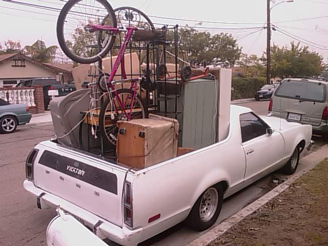White Ford Ranchero at curb, bed loaded with furniture, boxes and a pink bicycle