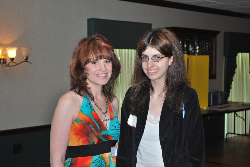 Two women smiling at the camera indoors. The woman on the left wears a colorful dress and necklace. The woman on the right wears glasses and a black jacket