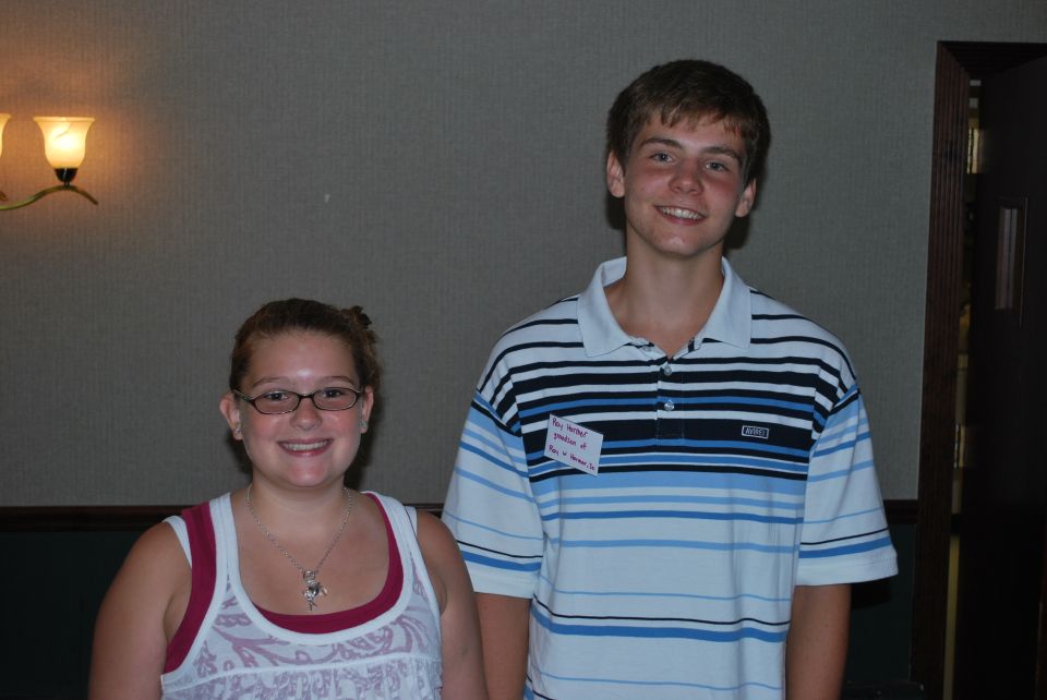 Two smiling young people, a girl with glasses and a boy in a striped shirt, stand indoors