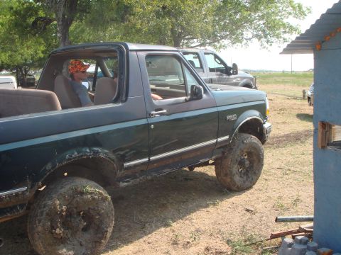 1992 Ford Bronco - A dark green 1992 Ford Bronco with large mud tires sits off-road next to a pickup truck.