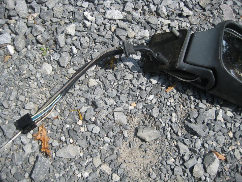 Broken side mirror assembly with exposed wires and connector, lying on gravel