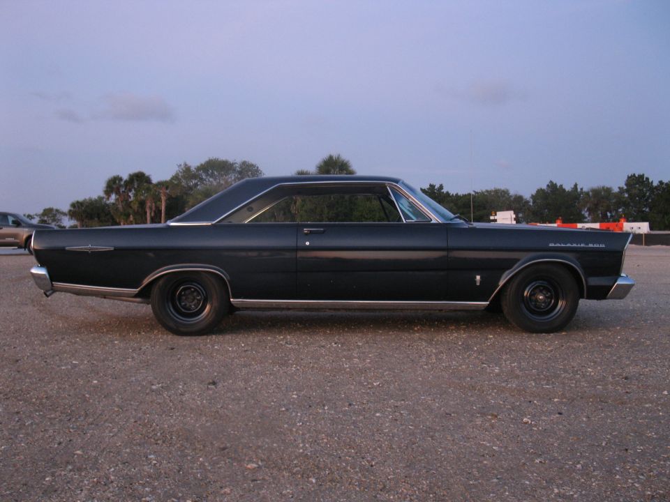 Dark blue Ford Galaxie 500 fastback hardtop side profile on gravel at dusk with black steel wheels