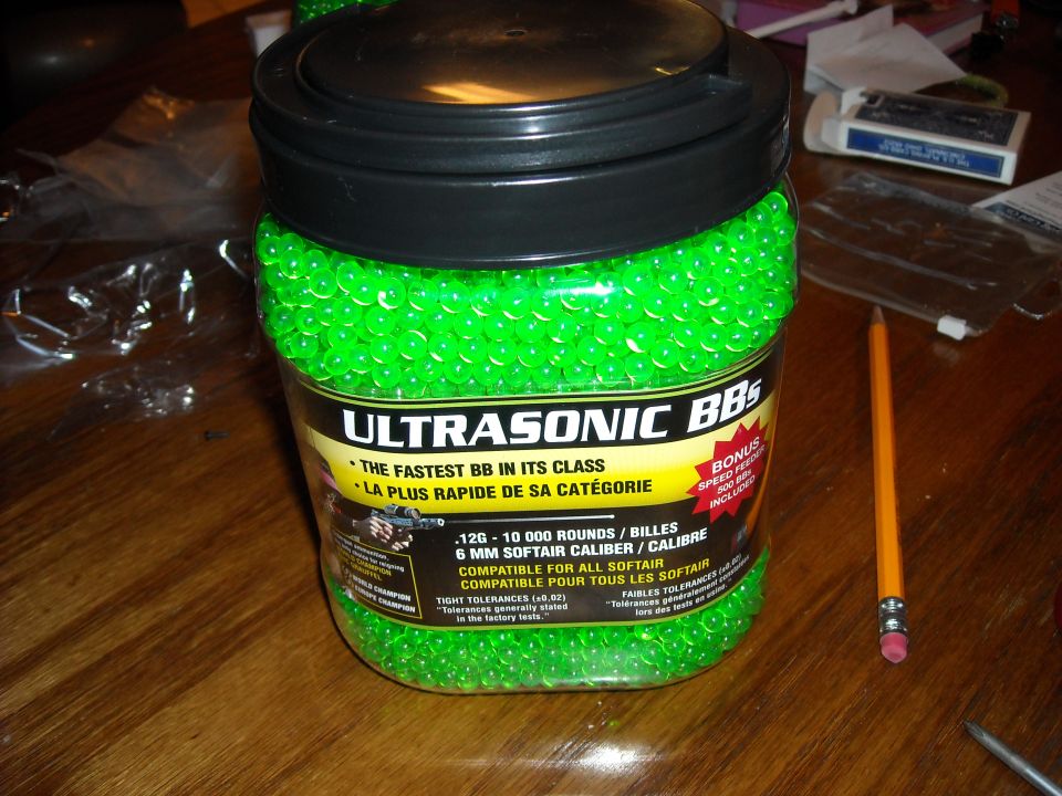 Jar of green Ultrasonic BBs, 10,000 rounds, 6mm softair caliber. Pencil and other items on wooden table