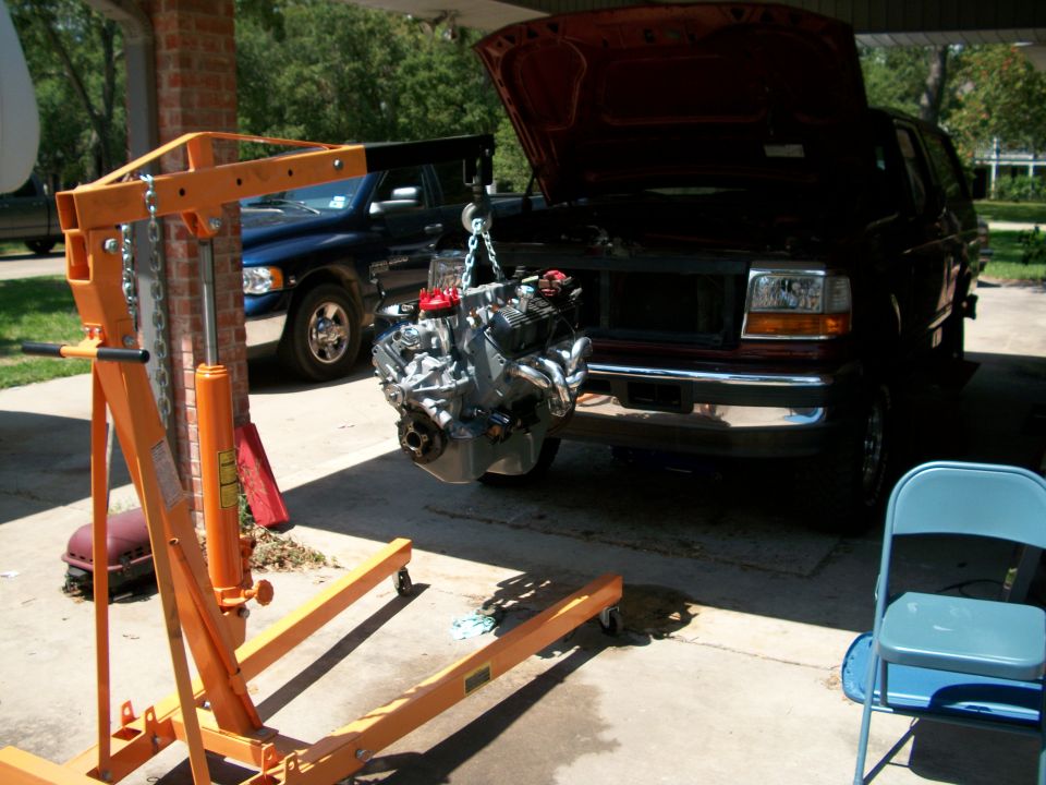 Rebuilt V8 engine hanging from an orange hoist beside a dark red Ford SUV with open hood