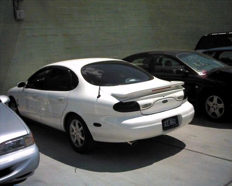 1990 Ford Taurus - A white sedan with a spoiler is parked next to a black car, with a license plate visible.