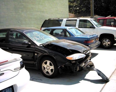 1990 Ford Taurus - A damaged black Ford Taurus is parked in a lot with other vehicles.