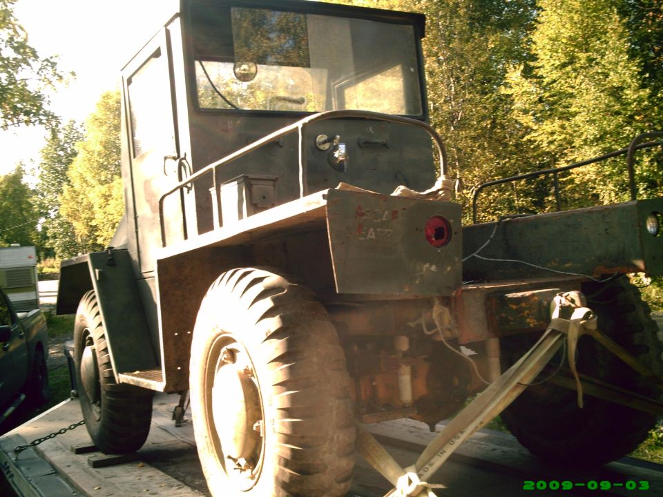 Rusty olive-drab military-style vehicle cab and chassis strapped to a flatbed trailer