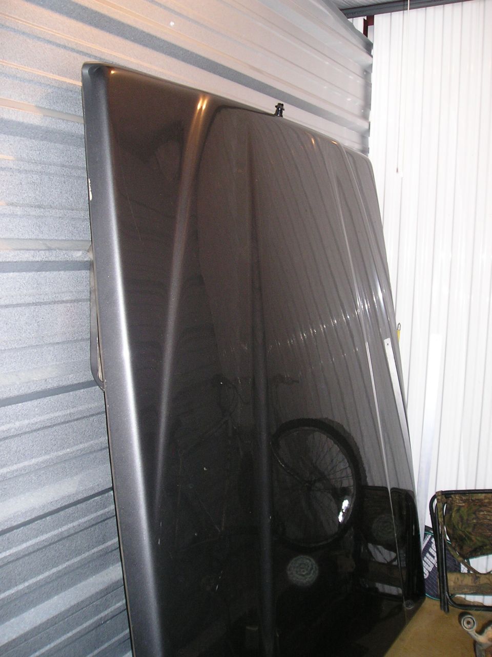 Dark colored truck hood leaning against a wall inside a storage unit. A fan is reflected in the paint