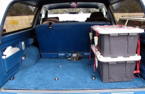 1984 Ford Bronco - The cargo area of an SUV shows blue carpeting and seats, with two storage bins stacked and strapped down.