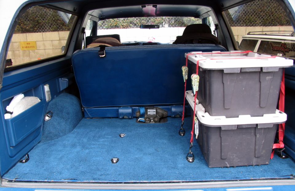 Interior cargo area with blue carpet, blue seats, and two stacked gray storage bins secured with red straps