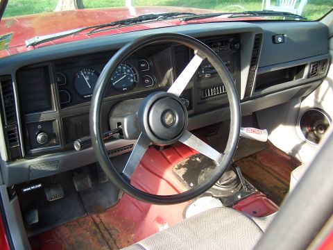 1991 Jeep Comanche - The interior of a red Jeep shows an aftermarket steering wheel and worn floor mats.