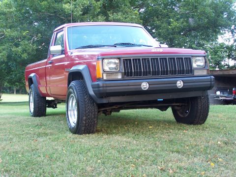 1991 Jeep Comanche - Red Jeep Comanche pickup parked on grass, three-quarter front view showing the seven-slot grille with 'Jeep' badge, black fender flares, ro…