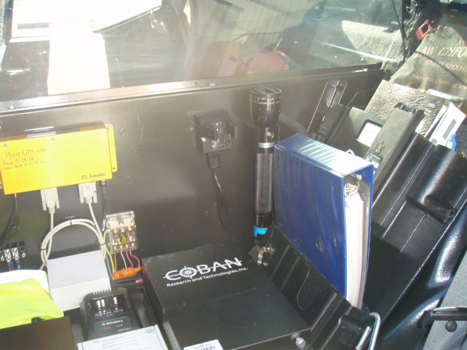 Police car interior with yellow GPS unit, flashlight, blue binder, and COBAN equipment