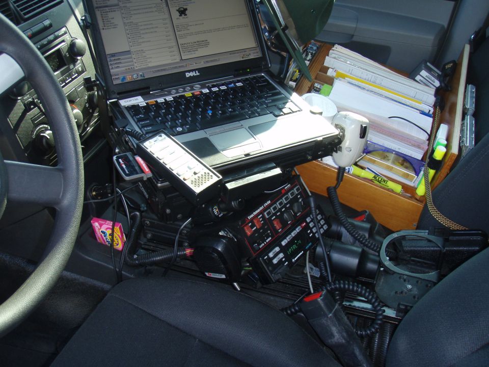 Interior of a police car showing a laptop, radio equipment, and a wooden organizer with papers