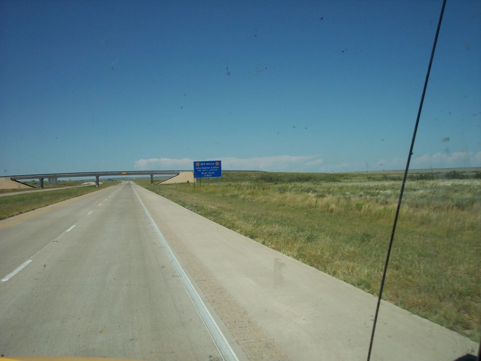 Highway scene with a blue sign indicating New Mexico, a bridge, and grassy plains under a clear blue sky
