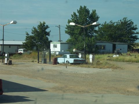 1960 Ford Falcon - A light blue sedan from the 1960s is parked at a rural gas station with trailers and trees in the background.