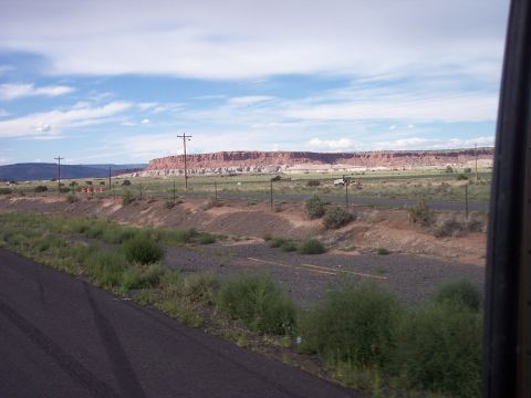 1960 Ford Falcon - A wide landscape shot shows distant vehicles on a road with a prominent red rock formation in the background under a partly cloudy sky.